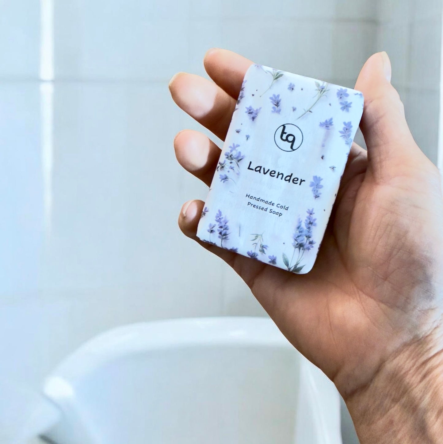 “Hand holding a Truequest lavender cold‑pressed soap wrapped in white paper with purple flower illustrations.