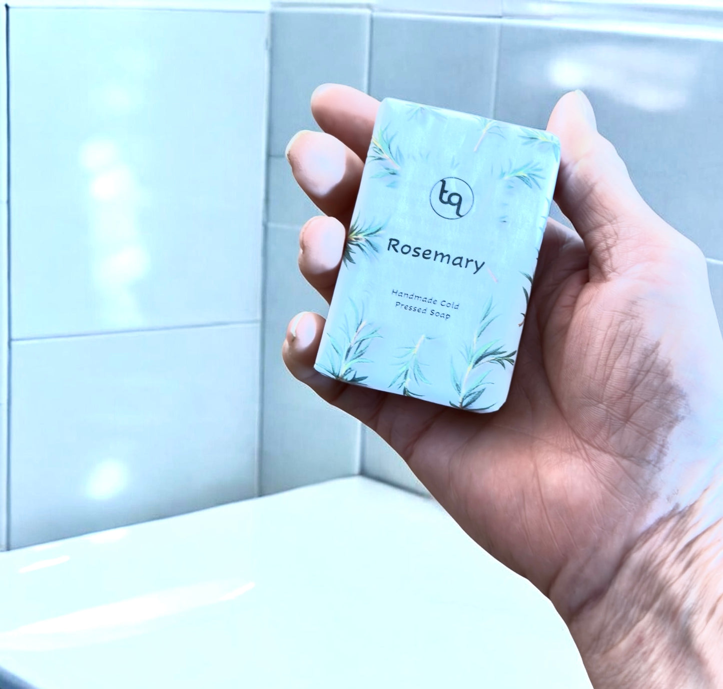 A hand holding a blue‑wrapped Truequest rosemary cold‑pressed soap bar with a subtle leaf pattern.