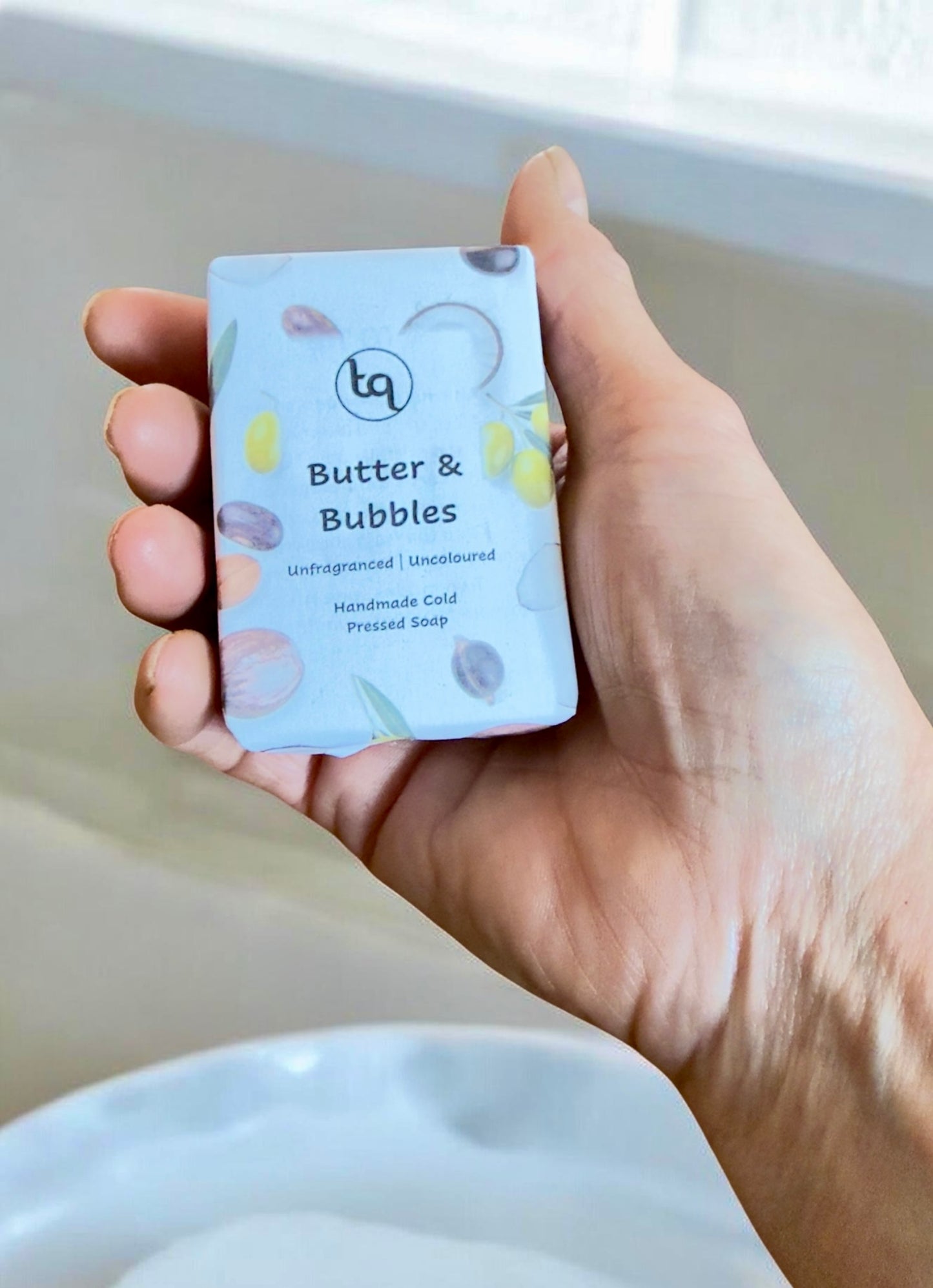 Hand holding a bar of Truequest soap labeled 'Butter & Bubbles' with a bathtub in the background.