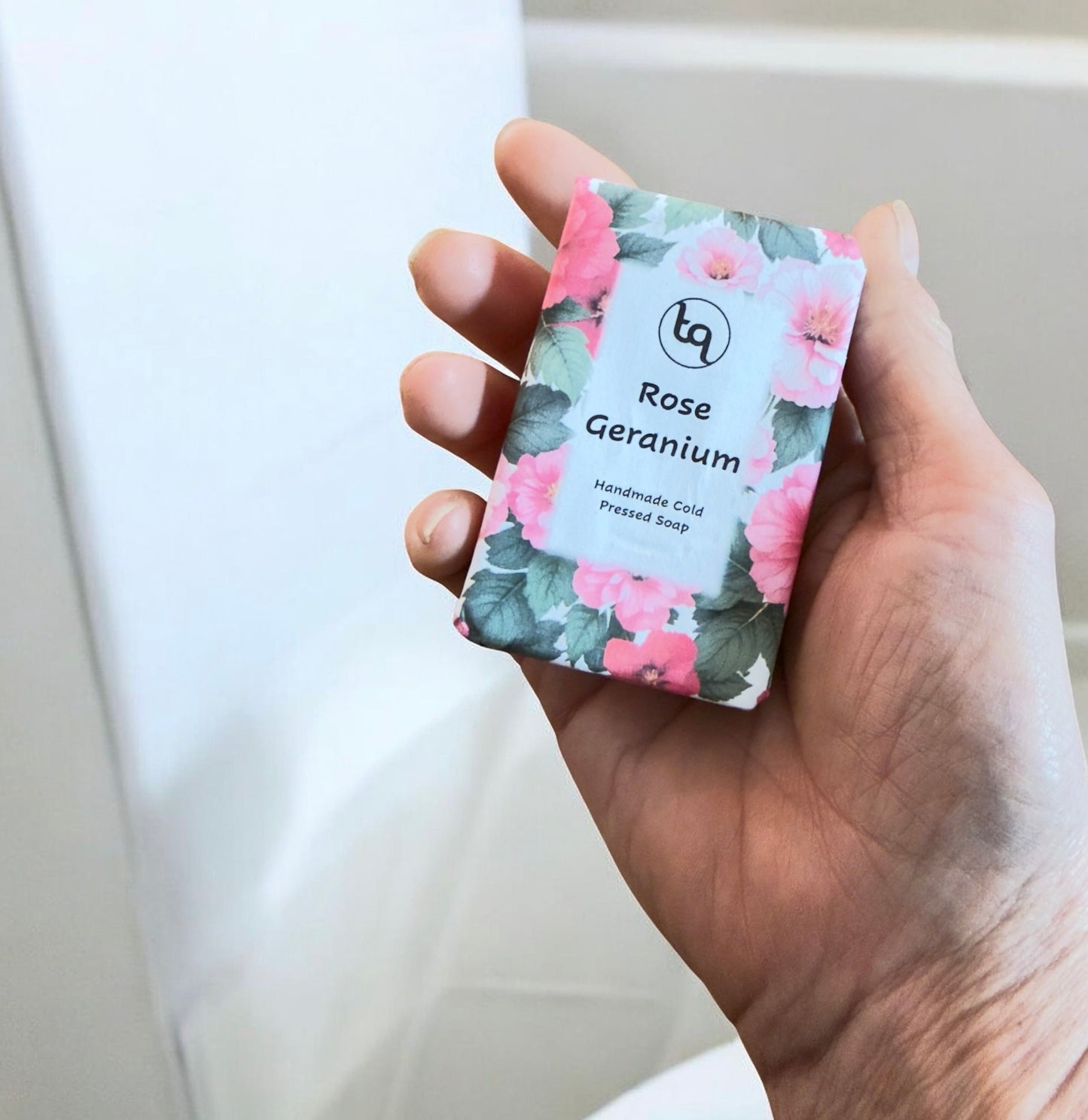 Hand holding a soap bar with floral packaging labeled 'Rose Geranium'.