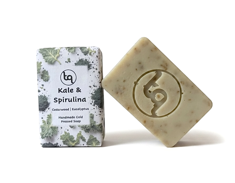 A product photo of two Truequest kale and spirulina cold-pressed soap bars, featuring one bar neatly enclosed in its paper packaging and a second unwrapped bar resting at a dynamic angle to show its deep green colour