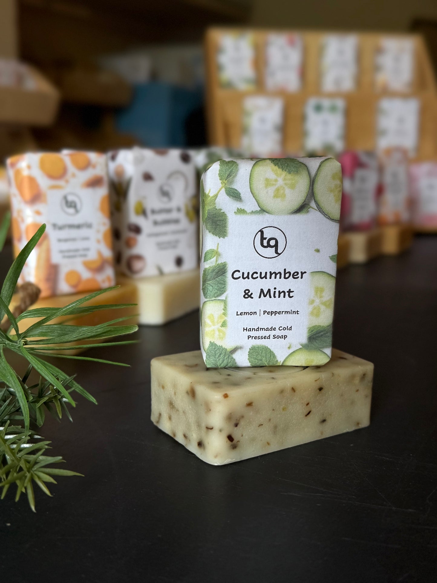Bar of soap with a 'Cucumber & Mint' label on a dark surface, surrounded by other soaps and packaging.
