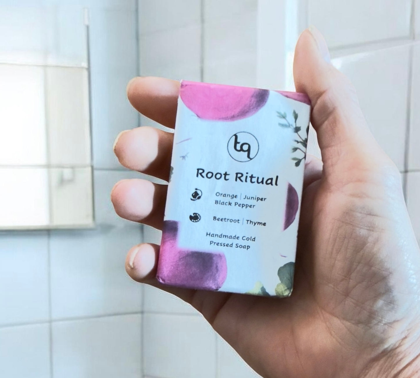 Hand holding a Truequest soap bar with 'Root Ritual' packaging against a tiled bathroom wall.