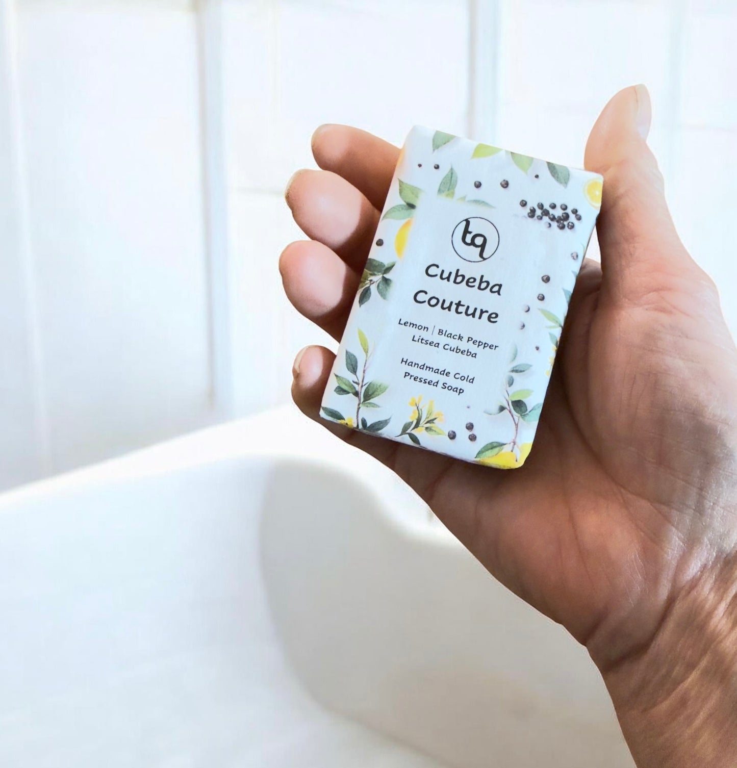Hand holding a Truequest Cubeba Couture soap wrapped in botanical paper with lemon slices, leaves and peppercorn illustrations.