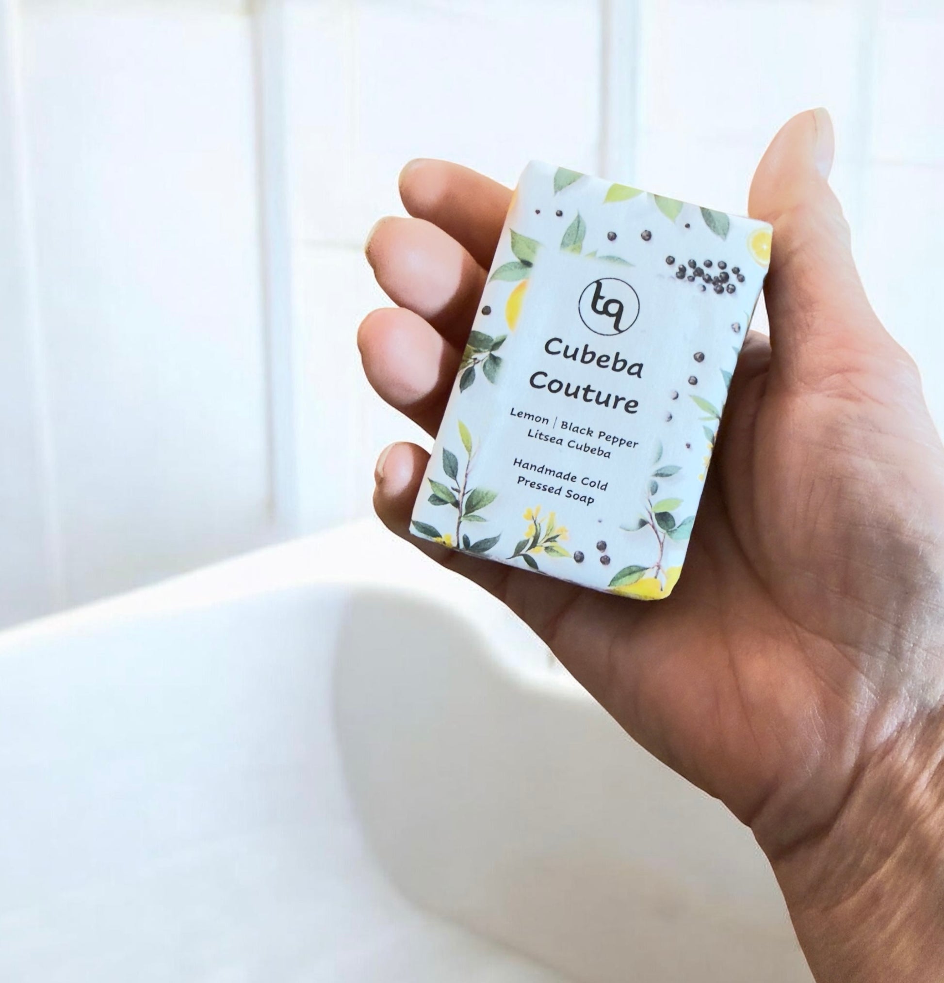 Hand holding a Truequest Cubeba Couture soap wrapped in botanical paper with lemon slices, leaves and peppercorn illustrations.