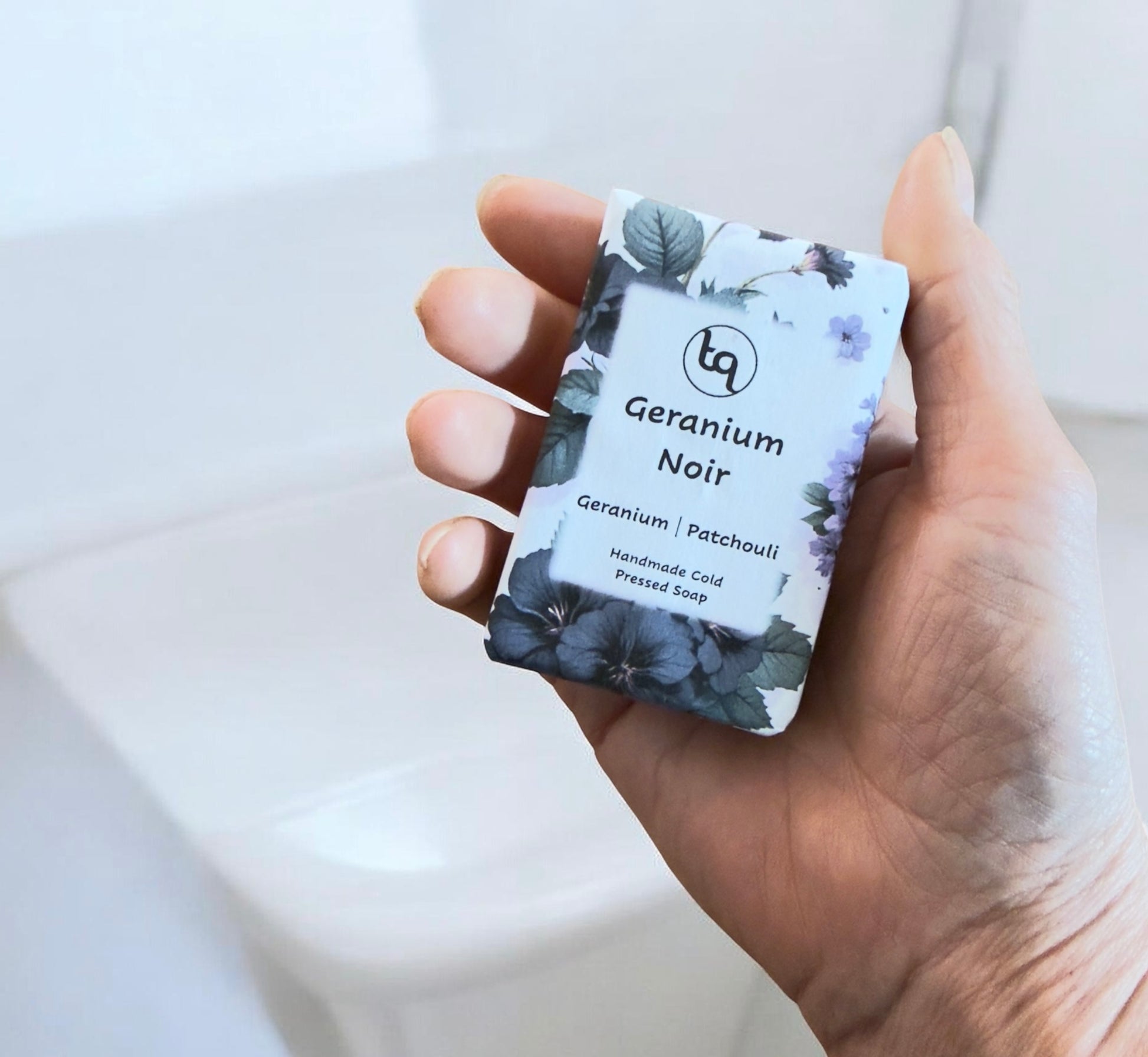 Hand holding a Truequest Geranium Noir soap wrapped in blue and purple floral paper.
