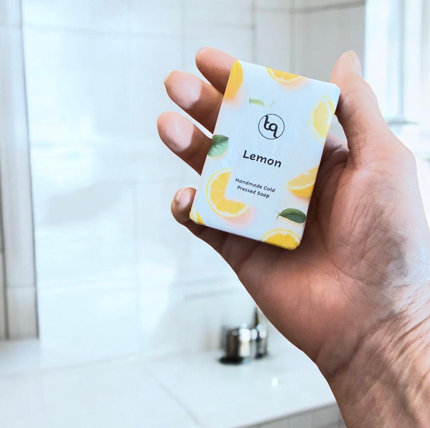 Hand holding a Truequest lemon cold‑pressed soap wrapped in white paper with yellow citrus slices and green leaves.