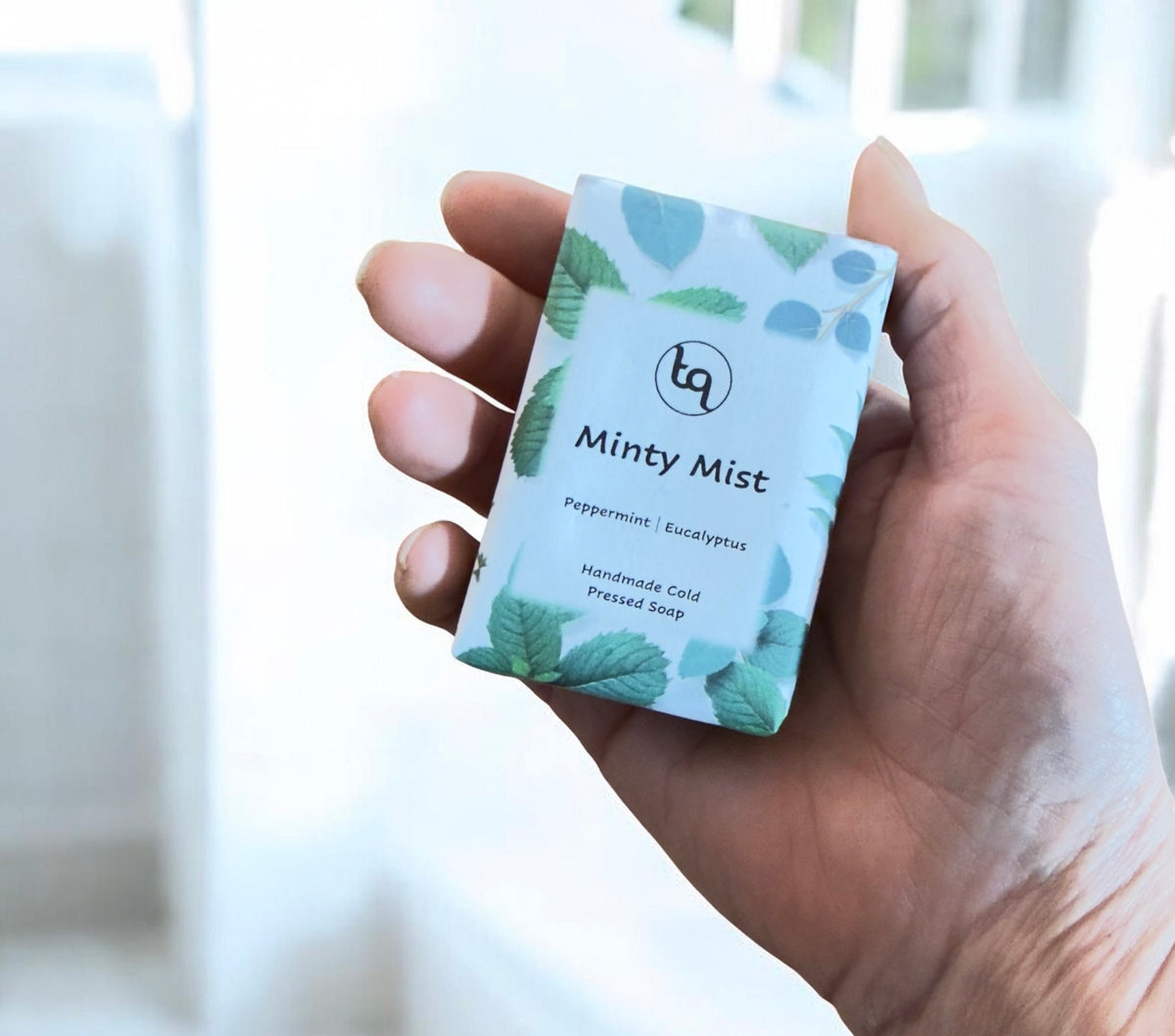 Hand holding a Truequest Minty Mist cold pressed soap wrapped in green mint leaf patterned paper.