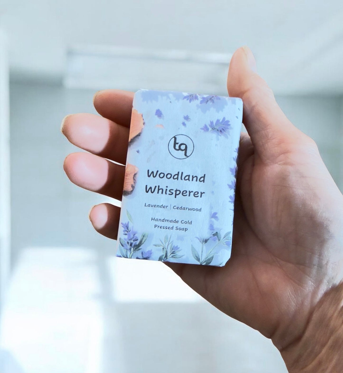 Hand holding a bar of soap labeled 'Woodland Whisperer'  lavender and cedarwood from Truequest soap with a blurred background